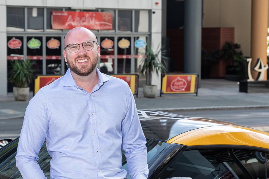 Stephen Cornish, a white man in a blue button up shirt, smiles at the camera on a city street.