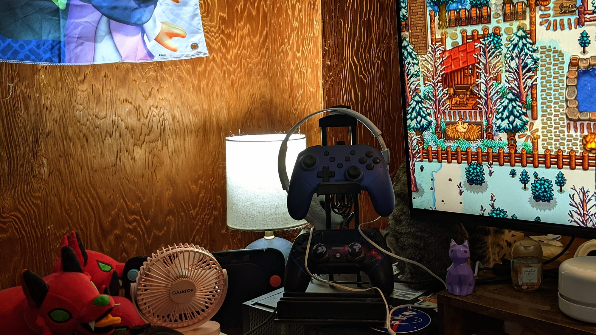 The corner of a desk by a wood panel wall. The desk is full of little trinkets and plush toys, with two purple space-themed controllers sitting on a sticker-covered computer case.