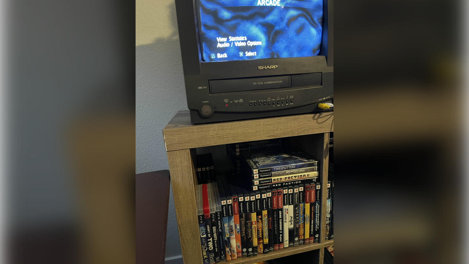 A CRTV with built in VCR sits above a shelf full of retro video games, largely for PlayStation 2.