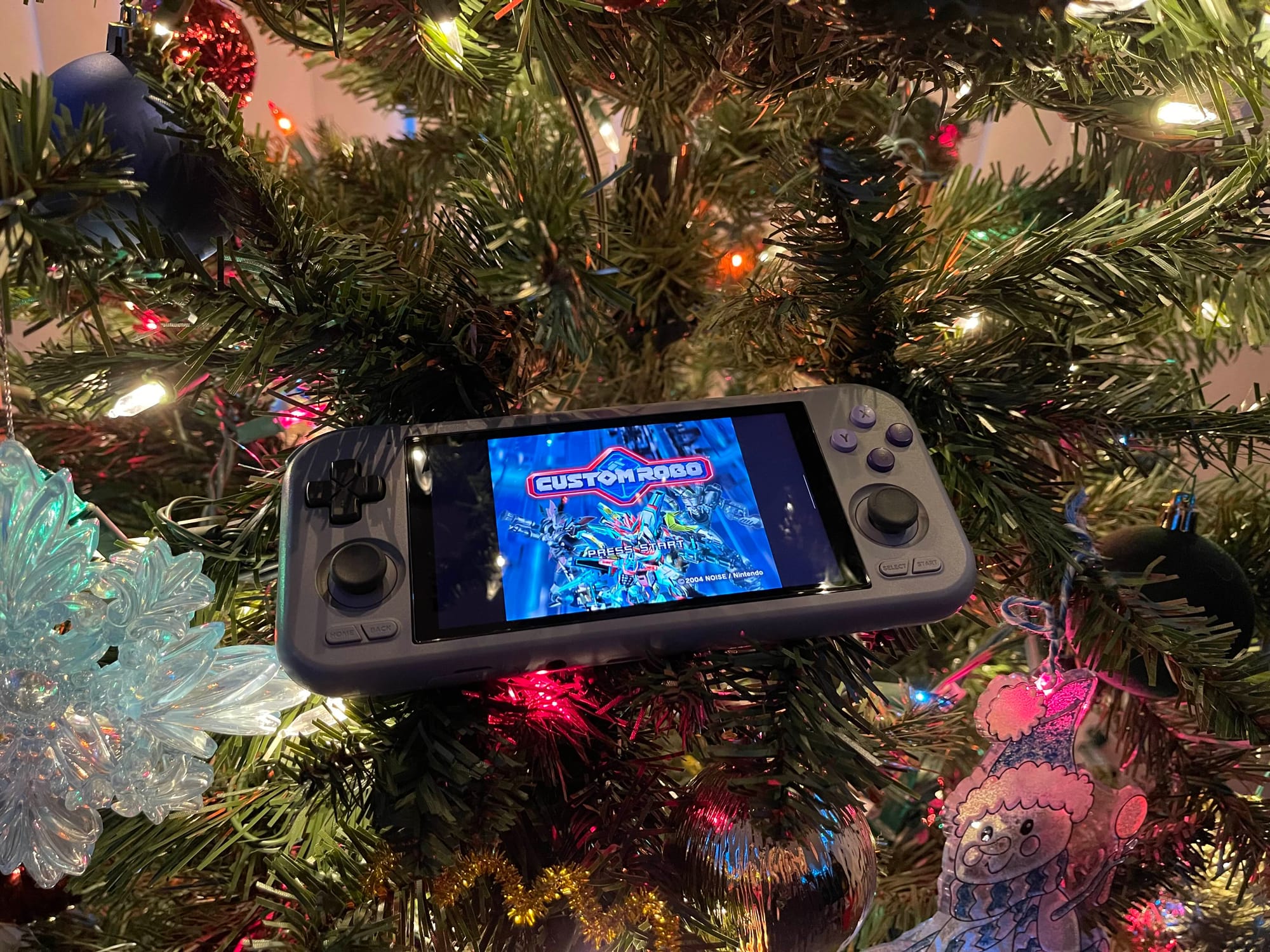 A handheld gaming console sits in a decorated Christmas tree lit with multicolor lights.