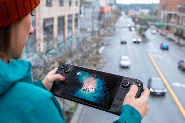 A woman in a blue hoodie plays Hollow Knight on a Steam Deck while overlooking a city street.