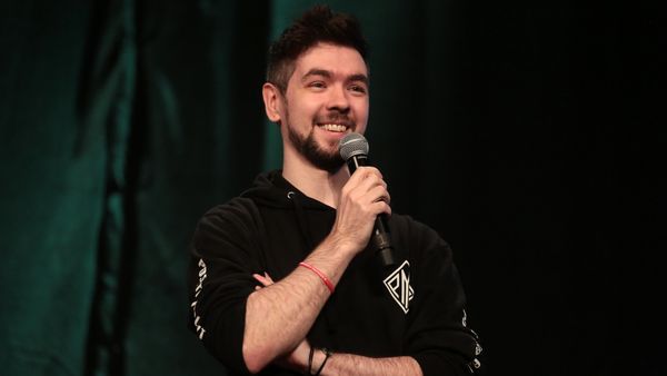 A white man with short brown hair and a short beard holds a microphone up and smiles at the audience behind the camera.